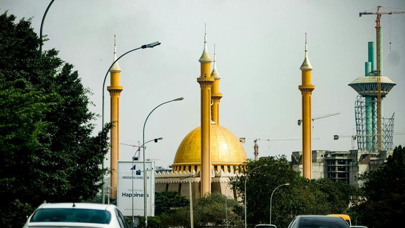 Cars on a tree-lined Abuja road with the golden dome and minarets of the National Mosque and construction cranes behind