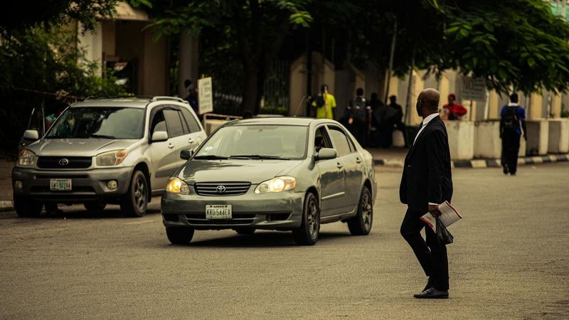 A man in a dark suit crossing a tree-lined Abuja street as cars wait beside him