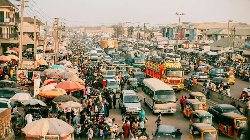 Busy Nigerian city road packed with cars, buses, keke tricycles and lorries beside crowded market stalls under umbrellas