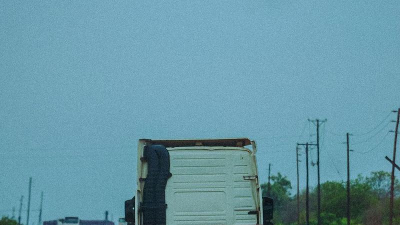 Rear view of a red flatbed lorry carrying a white truck cab along a road lined with electricity poles