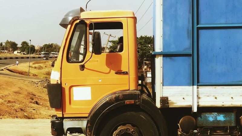 An orange-cab truck with a blue box body parked on a concrete yard beside a highway