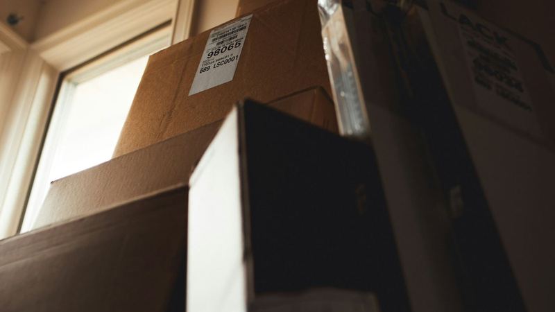 Stack of labelled cardboard boxes piled high beside a window in a dimly lit room
