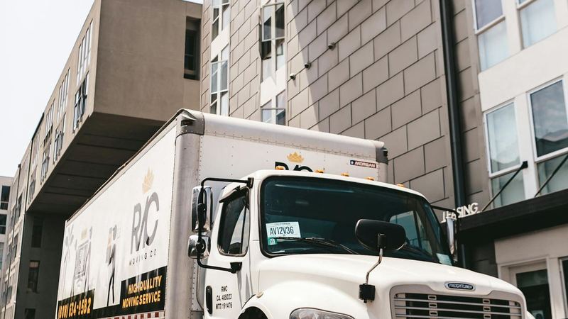 White box moving truck parked in front of a modern multi-storey apartment building on a bright day