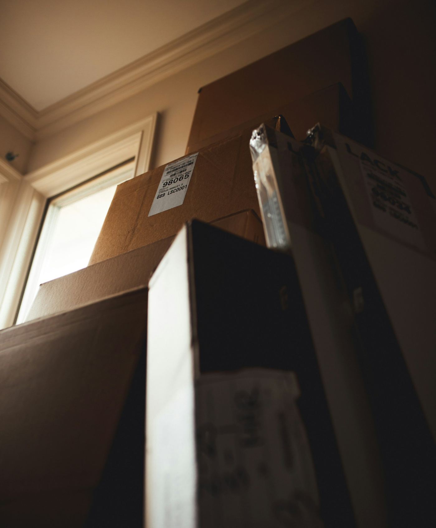 Stack of labelled cardboard boxes piled high beside a window in a dimly lit room